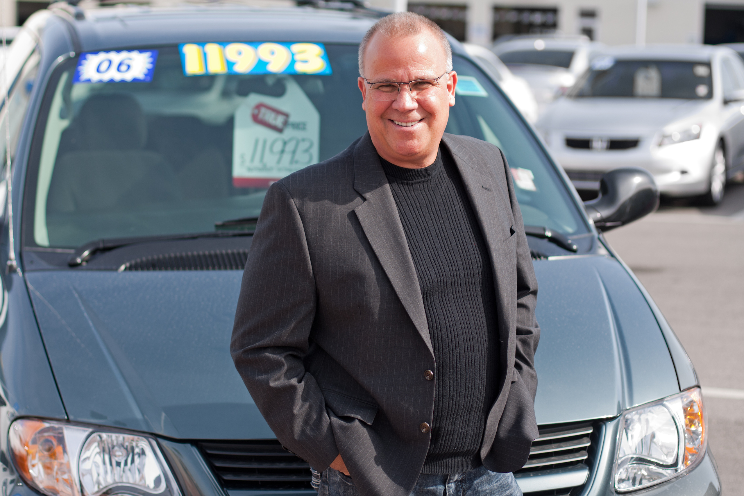 Happy car dealer leaning on the hood of a car for sale